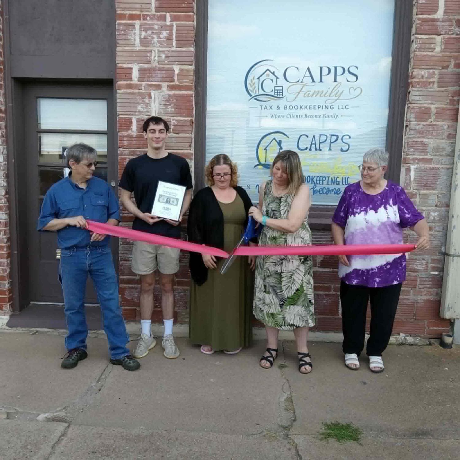 Ribbon cutting outside the Capps Family Tax and Bookkeeping office in downtown Lyons, Kansas. Left to right: John Capps, CEO Philip Capps, Lyons Chamber of Commerce director Ariah Allensworth, office manager Sherri Capps cutting the pink ribbon with oversized scissors, and receptionist Margaret. Philip holds a Lyons Chamber of Commerce congratulations plaque.